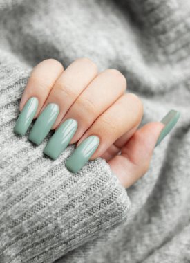 Güzel manikürlü, yeşil tırnaklı şık bir el yumuşak gri bir kazağın üzerine nazikçe yerleştirilir, rahat bir ortamda şık bir aksan sergiler..