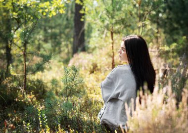 Genç bir kadın, yaprakların arasından süzülen uzun ağaçlar ve güneş ışığı ile çevrili yemyeşil çimlerin arasında huzur içinde oturup doğanın huzur verici varlığının tadını çıkarıyor..