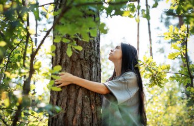 Bir kadın canlı bir ormanda duruyor, sevgiyle bir ağacı kucaklıyor. Güneş ışığı yaprakların arasından süzülür ve dingin atmosfere sıcaklık katar..