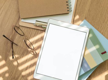 Boş defterler, okuma gözlükleri ve kırtasiye malzemeleriyle çevrili ahşap bir masanın üzerinde duran tablet çalışma alanını hafifçe aydınlatıyor..