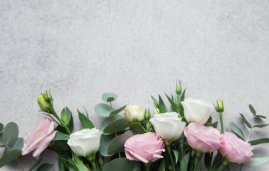 Yeşil okaliptüs yapraklarının yanında, soluk gri bir yüzeyde, zarif pembe ve beyaz güllerin doğal güzelliklerini ve zarafetlerini vurgulayan zarif bir düzenleme uzanıyor..