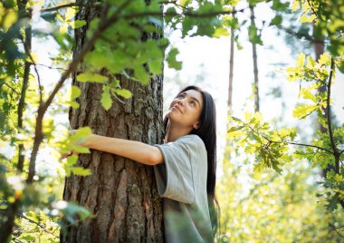 Bir kadın canlı bir ormanda duruyor, sevgiyle bir ağacı kucaklıyor. Güneş ışığı yaprakların arasından süzülür ve dingin atmosfere sıcaklık katar..