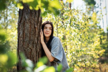 Genç bir kız ormanda bir ağaca sarılıyor. Stresi azaltmak ve doğayla bağ kurmak için ağaçlara sarılıp dokunmak..