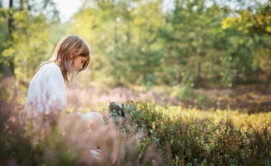 Renkli kır çiçekleri ve yeşilliklerle çevrili bir çocuk yerde dinlenir. Güneş ağaçların arasından süzülüp ormanda sakin bir atmosfer yaratıyor..