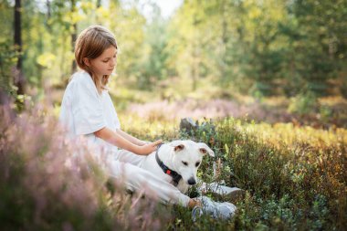 Genç bir kız ormanda huzurlu bir anın tadını çıkarıyor, beyaz köpeğini severek okşuyor, onlar yemyeşil yemyeşil bir ortamda dinlenirken..