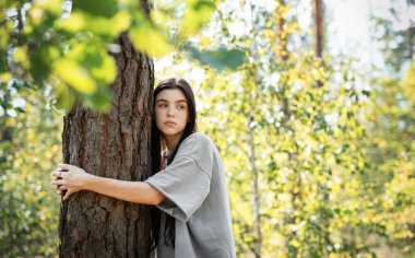 Bir kadın ağaca yaslanmış, güneşli bir ormanda canlı yeşil yapraklarla çevrili, dingin bir ifadeyle yukarıya bakıyor..