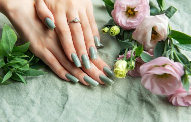 Güzel manikürlü ve yeşil tırnaklı bir çift el zarif pembe ve beyaz çiçeklerle çevrili dokulu bir kumaşa dayanır, huzurlu ve şık bir anı yakalar..