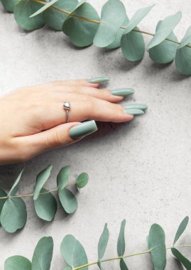 A hand with beautifully manicured green nails is gently placed on a light gray surface, surrounded by delicate eucalyptus leaves, creating a serene atmosphere. clipart