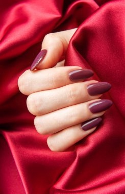 Manikürlü bir el, canlı kırmızı saten kumaşa karşı derin bordo çiviler sergiliyor. Samimi bir ortamda zarafet ve stili vurguluyor..