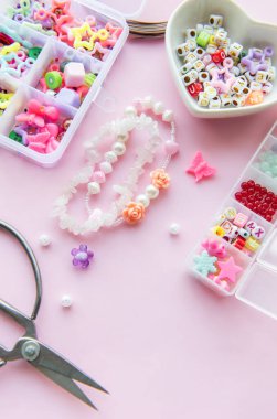 Renkli boncukların, tılsımların ve makasların pastel pembe yüzey üzerindeki yaratıcı dizilimi eşsiz mücevher yapma sürecini gözler önüne seriyor. Zarif çiçekli tasarımlar vurgulanır.