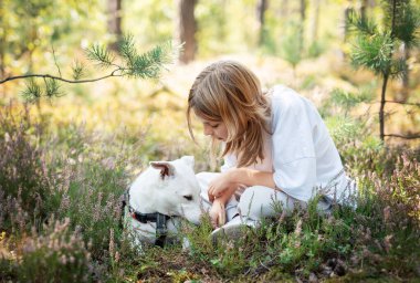 Bir çocuk yemyeşil bir ormanda oturuyor, renkli çiçekler ve yeşillikler arasında dost bir köpeği nazikçe okşuyor, doğada huzurlu bir anın tadını çıkarıyor..