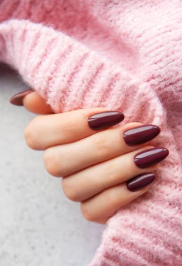 Bir kadının elinin yakın plan çekimi, koyu kırmızı badem şekilli çivileri zarif bir şekilde pembe bir kazağa sarıp nötr bir zemine yapıştırıyor..