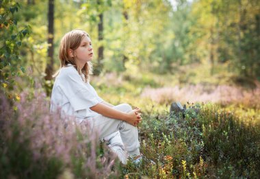 Uzun saçlı bir çocuk, verimli yeşilliklerin ve renkli bitkilerin arasında güneşli bir günde ormanın sessizliğinin tadını çıkararak huzur içinde oturur..