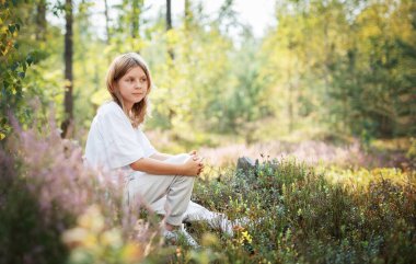Renkli kır çiçekleri ve yeşilliklerle çevrili bir çocuk yerde dinlenir. Güneş ağaçların arasından süzülüp ormanda sakin bir atmosfer yaratıyor..
