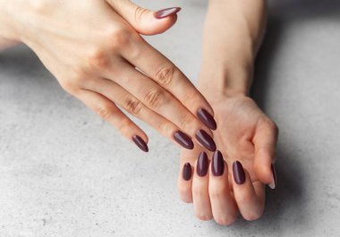 Bir çift el zarif bir şekilde uzun, badem şekilli, zengin bir kestane rengi ile boyanmış, pürüzsüz gri bir yüzey üzerinde dinlenen, zarafet ve stili vurgulayan çiviler sergiliyor..