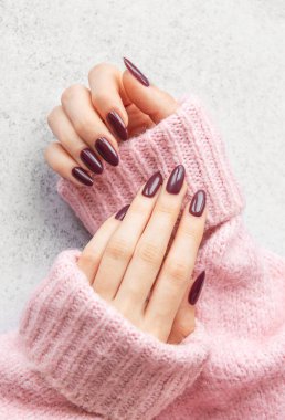 Güzelce hazırlanmış kestane rengi tırnaklar rahat pembe bir kazağın üzerinde dinlenirken gösteriliyor, ince gri bir arka plana karşı kontrastı vurguluyor, şık ve davetkar bir görünüm yaratıyor..
