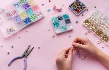 Çeşitli boncuklar ve zanaat araçlarıyla çevrelenmiş, yumuşak pembe bir zemin üzerine kurulmuş, kolye yapmak için eller ustalıkla dizilmiş..