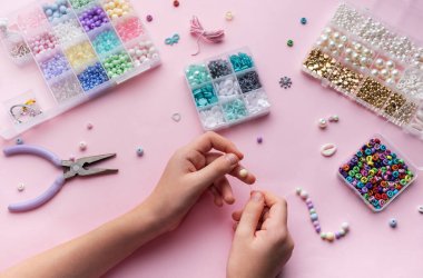 Bir kişi, boncuk, alet ve materyal konteynerleri ile çevrili yumuşak pembe bir yüzey üzerinde boncuk mücevherler monte ediyor ve yaratıcı bir zanaat süreci sergiliyor..