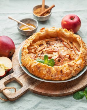 Yeni pişmiş bir elmalı turta, badem ve nane ile süslenmiş, ahşap bir tahtanın üzerinde oturur, yanında kırmızı elmalar ve küçük bir bal kasesi, sıcak bir atmosferi davet eder..