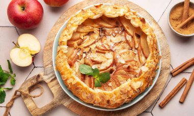 Güzel pişmiş bir elmalı turta, taze elma ve tarçın çubuklarıyla çevrili ahşap bir tahtanın üzerinde oturur ve sıcak ve davetkar bir mutfak atmosferi yaratır..