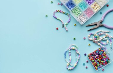 Yumuşak mavi arkaplan üzerindeki boncuklar, aletler ve bitmiş bileziklerden oluşan canlı bir düzenleme çeşitli renk ve tasarımlarla mücevher yapma sanatını gözler önüne seriyor..