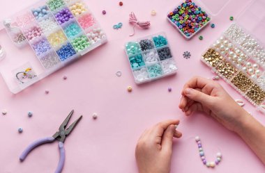 Çeşitli boncuklar ve zanaat araçlarıyla çevrelenmiş, yumuşak pembe bir zemin üzerine kurulmuş, kolye yapmak için eller ustalıkla dizilmiş..