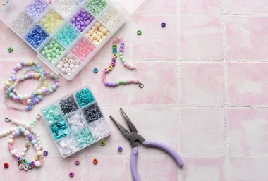 Kişiselleştirilmiş bilezikler yapmak için pembe bir zemine yerleştirilmiş çeşitli renkli boncuklar, mektup boncukları ve mücevher penseleri.