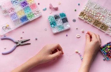 Bir kişi, boncuk, alet ve materyal konteynerleri ile çevrili yumuşak pembe bir yüzey üzerinde boncuk mücevherler monte ediyor ve yaratıcı bir zanaat süreci sergiliyor..