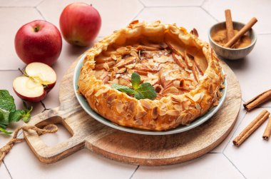 Güzel pişmiş bir elmalı turta, taze elma ve tarçın çubuklarıyla çevrili ahşap bir tahtanın üzerinde oturur ve sıcak ve davetkar bir mutfak atmosferi yaratır..