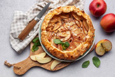 Badem gevreği ve nane ile süslenmiş taze pişmiş elma galetesi, taze elmalar ve bir bıçakla servis edilen bir masada.