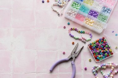 Renkli boncuklar, harf boncukları, ip, pense ve pense içeren el işi malzemeler pembe bir fayans yüzeyinde, mücevher yapımına hazır.