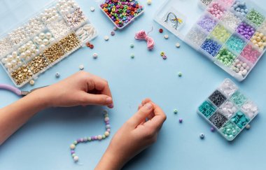 Bir kişi parlak bir el sanatları masasında çeşitli renkli boncuklar ve aletlerle çevrili bir diziye ustalıkla iplik geçirir..