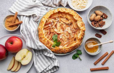 Badem ve bal ile süslenmiş güzel sunulmuş bir elmalı tart kırsal bir masada oturur. Etrafında taze elmalar, tarçın çubukları ve fındık kaseleri var. Sıcak, davetkar bir atmosfer yaratıyorlar..