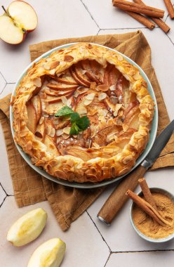 Badem yaprakları, tarçın çubukları ve nane yapraklarıyla süslenmiş taze pişmiş elma galetesi beyaz fayanslı bir masada oturuyor.