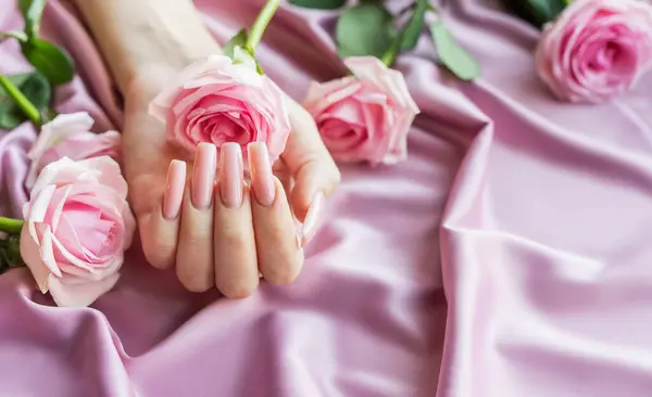 Woman\'s hand with fashionable pink manicure holding pink rose flowers on pink silk fabric background