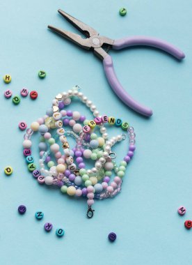 Yumuşak mavi arkaplan üzerindeki boncuklar, aletler ve bitmiş bileziklerden oluşan canlı bir düzenleme çeşitli renk ve tasarımlarla mücevher yapma sanatını gözler önüne seriyor..