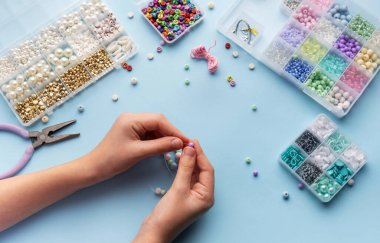 Bir kişi parlak bir el sanatları masasında çeşitli renkli boncuklar ve aletlerle çevrili bir diziye ustalıkla iplik geçirir..