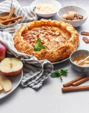 Güzel düzenlenmiş bir elmalı turtada badem, bal ve tarçınla kaplı pullu bir kabuk bulunur. Taze elma dilimleri ve çeşitli fındıklar tabağın etrafını sararak ılık bir sonbahar atmosferi yaratıyor..