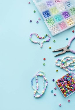 Renkli boncuklar, harf boncukları, pense ve bitmiş bilezikler açık mavi bir yüzey üzerinde, el yapımı için hazırlar.