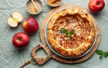 Yeni pişmiş bir elmalı turta, badem ve nane ile süslenmiş, ahşap bir tahtanın üzerinde oturur, yanında kırmızı elmalar ve küçük bir bal kasesi, sıcak bir atmosferi davet eder..