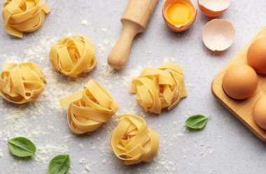 Pişmemiş fettucine makarna yuvalar, fesleğen yaprakları, yumurta, un ve merdane mutfak masasına dizilir ve kırsal bir mutfak sahnesi oluşturulur.