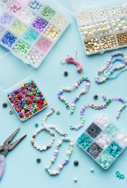 Yumuşak mavi arkaplan üzerindeki boncuklar, aletler ve bitmiş bileziklerden oluşan canlı bir düzenleme çeşitli renk ve tasarımlarla mücevher yapma sanatını gözler önüne seriyor..