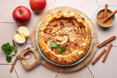 Güzel pişmiş bir elmalı turta, taze elma ve tarçın çubuklarıyla çevrili ahşap bir tahtanın üzerinde oturur ve sıcak ve davetkar bir mutfak atmosferi yaratır..