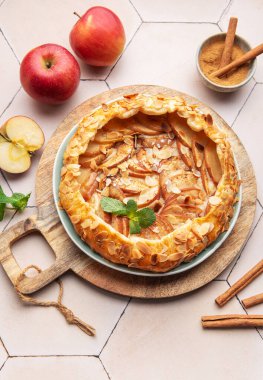 Güzel pişmiş bir elmalı turta, taze elma ve tarçın çubuklarıyla çevrili ahşap bir tahtanın üzerinde oturur ve sıcak ve davetkar bir mutfak atmosferi yaratır..