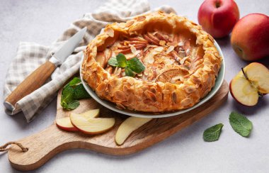 Badem gevreği ve nane ile süslenmiş taze pişmiş elma galetesi, taze elmalar ve bir bıçakla servis edilen bir masada.