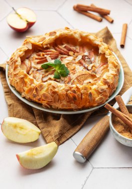 Badem yaprakları, tarçın çubukları ve nane yapraklarıyla süslenmiş taze pişmiş elma galetesi beyaz fayanslı bir masada oturuyor.