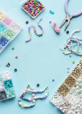 Yumuşak mavi arkaplan üzerindeki boncuklar, aletler ve bitmiş bileziklerden oluşan canlı bir düzenleme çeşitli renk ve tasarımlarla mücevher yapma sanatını gözler önüne seriyor..