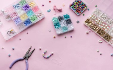Çalışma alanı bir dizi renkli boncuk, pense ve iplerle dolu, hepsi de yumuşak pembe arka planda. Ögeler, çeşitli şekil ve boyutlarda sergilenen temiz kaplarda düzenlenir..