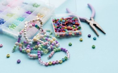 Renkli boncuklar, harf boncukları, pense ve bitmiş bilezikler açık mavi bir yüzey üzerinde, el yapımı için hazırlar.