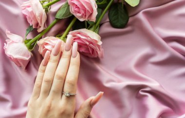 Pembe manikürlü ve nişan yüzüklü bir kadın eli ipek kumaşın üzerinde pembe güllerin yanında yatıyor.